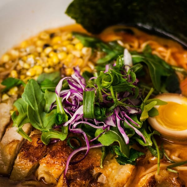 Close-up of a ramen bowl with chicken, corn, soft-boiled egg, greens, and red cabbage.