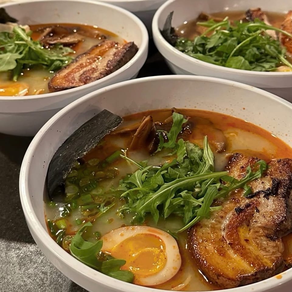 Three bowls of ramen topped with roasted pork, soft-boiled eggs, greens, mushrooms, scallions, and seaweed.