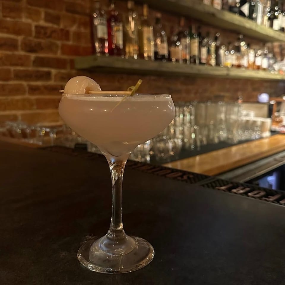Creamy pale cocktail garnished with a citrus twist in a coupe glass at a warmly lit bar with brick walls and liquor shelves.