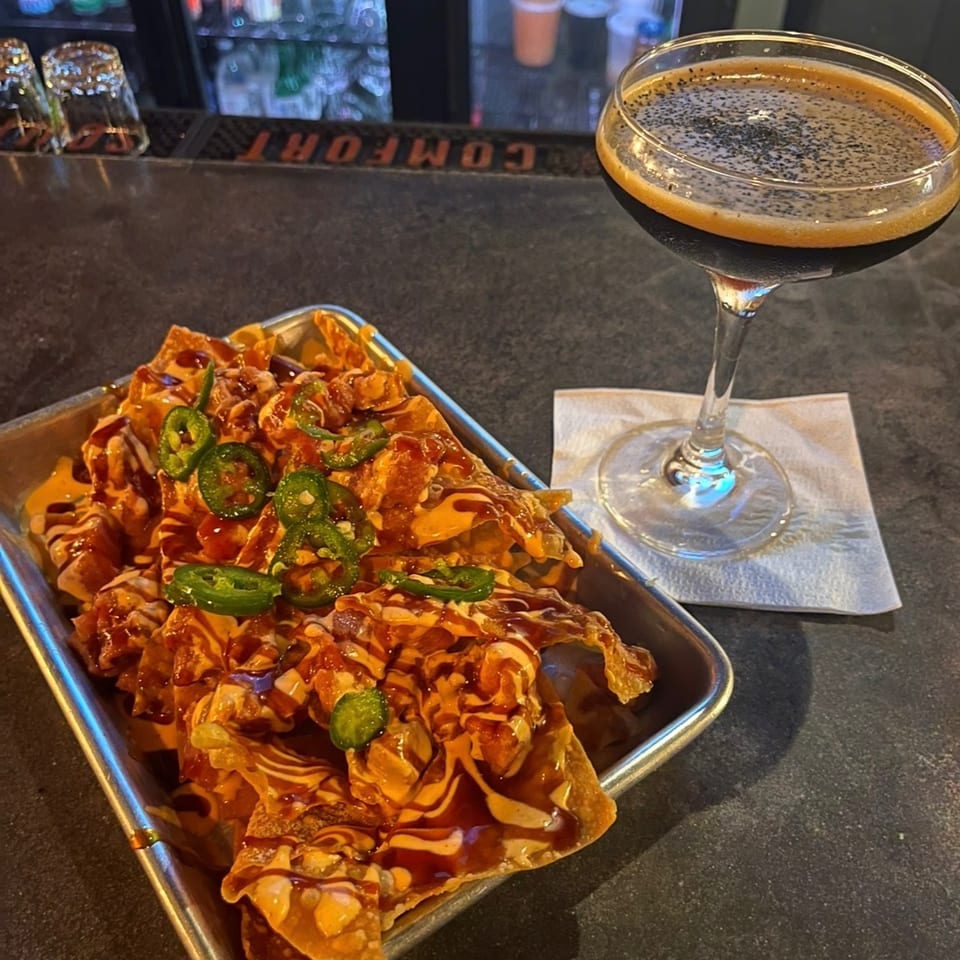 Loaded nachos topped with jalapeños, cheese, and sauces beside a foamy cocktail on a restaurant bar.