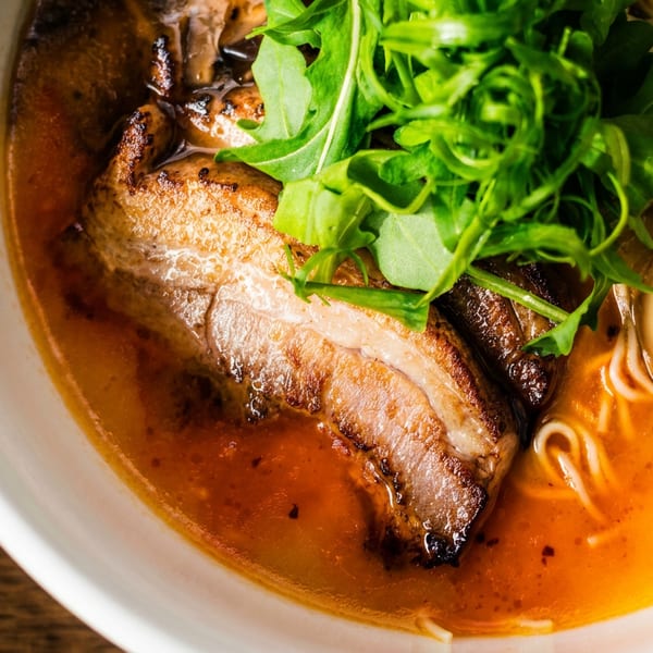 Close-up of a ramen bowl with pork belly, a soft-boiled egg, noodles, and arugula in a rich broth.