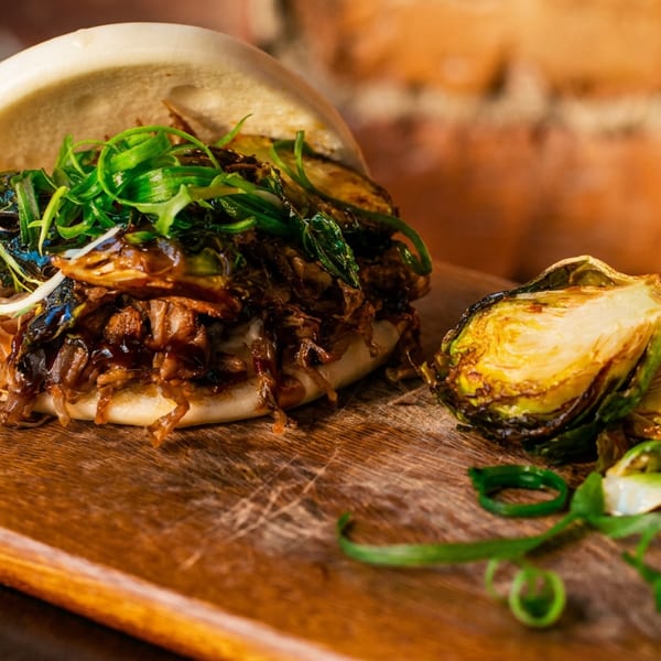 A bao bun filled with shredded meat and greens, served with roasted Brussels sprouts on a wooden board.