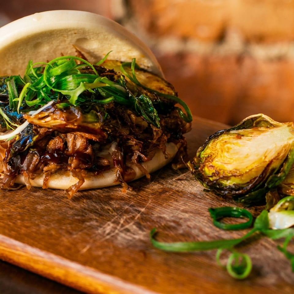 A bao bun filled with shredded meat and greens, served with roasted Brussels sprouts on a wooden board.