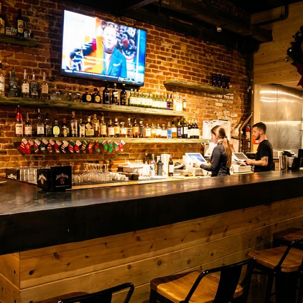 A rustic bar with brick walls, a TV, and shelves of liquor, with bartenders working.