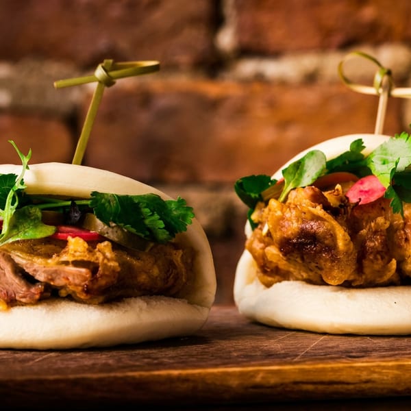 Two gourmet bao buns with meat, fresh herbs, and pickled vegetables on a wooden board.