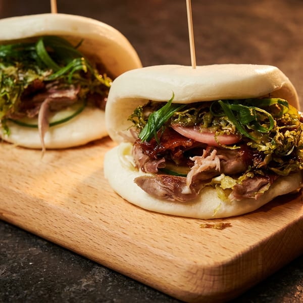 Two bao buns filled with meat, vegetables, and sauce, served on a wooden board.