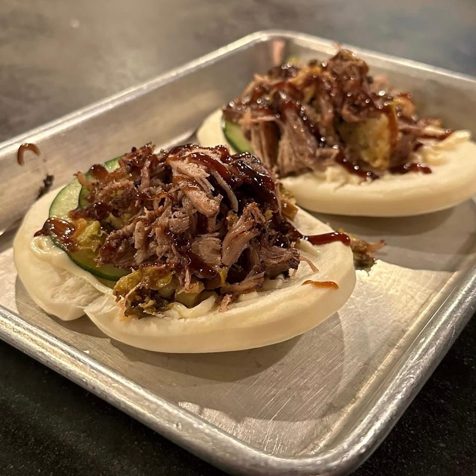 Two steamed bao buns filled with shredded meat, cucumber, vegetables, and glossy brown sauce on a metal tray.