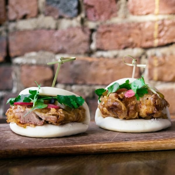 Two steamed bao buns filled with crispy meat, fresh herbs, and pickled radishes on a wooden board against a brick wall.